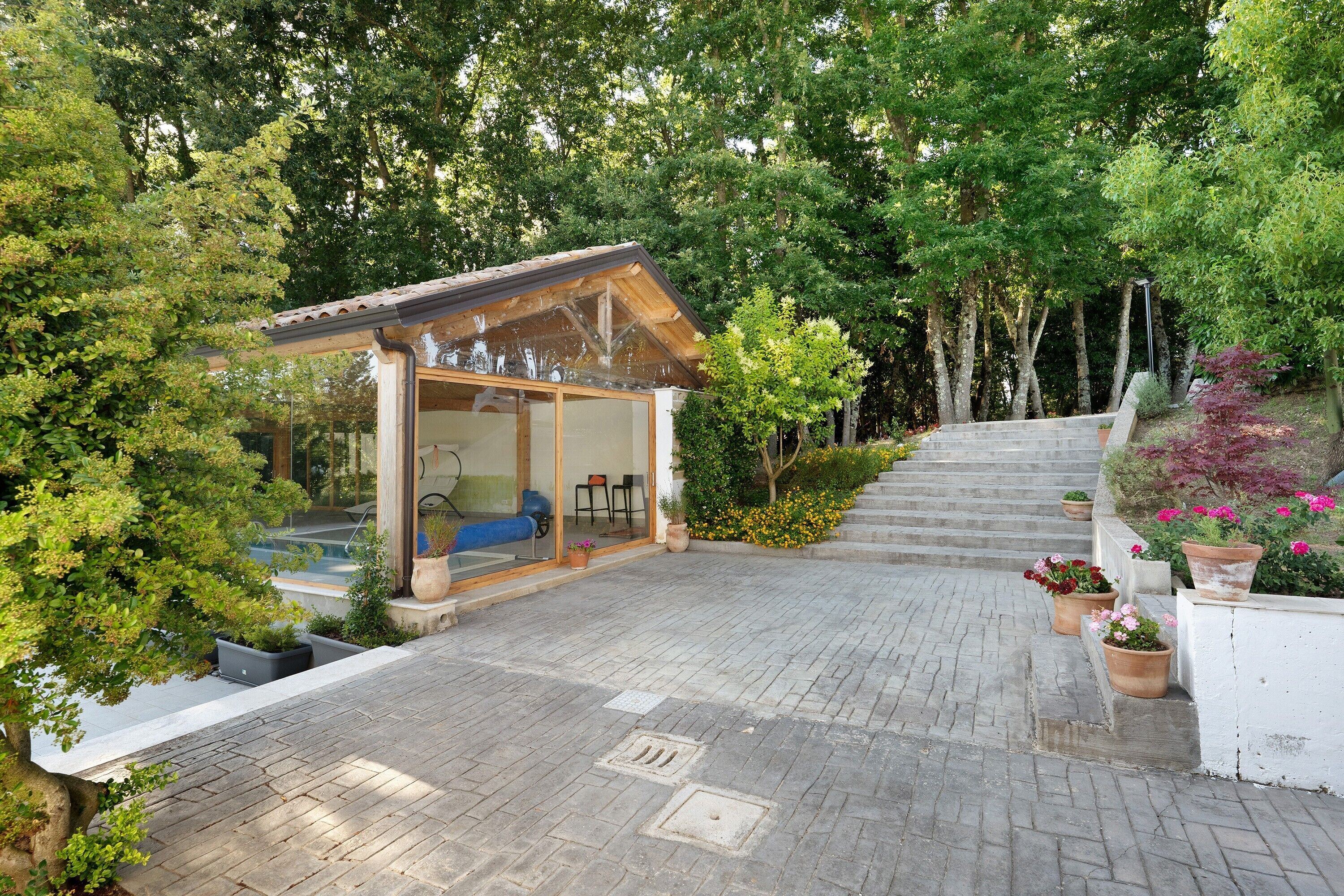 Un espace extérieur aménagé, abrité et doté de sièges, offrant une vue sur une piscine, le tout entouré d’arbres et de plantes en pot.