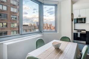 A modern kitchen with a stove, microwave, and cabinets. There is a dining area with a table and chairs, and a window with a city view. - NYC Luxe Rentals  (New York)