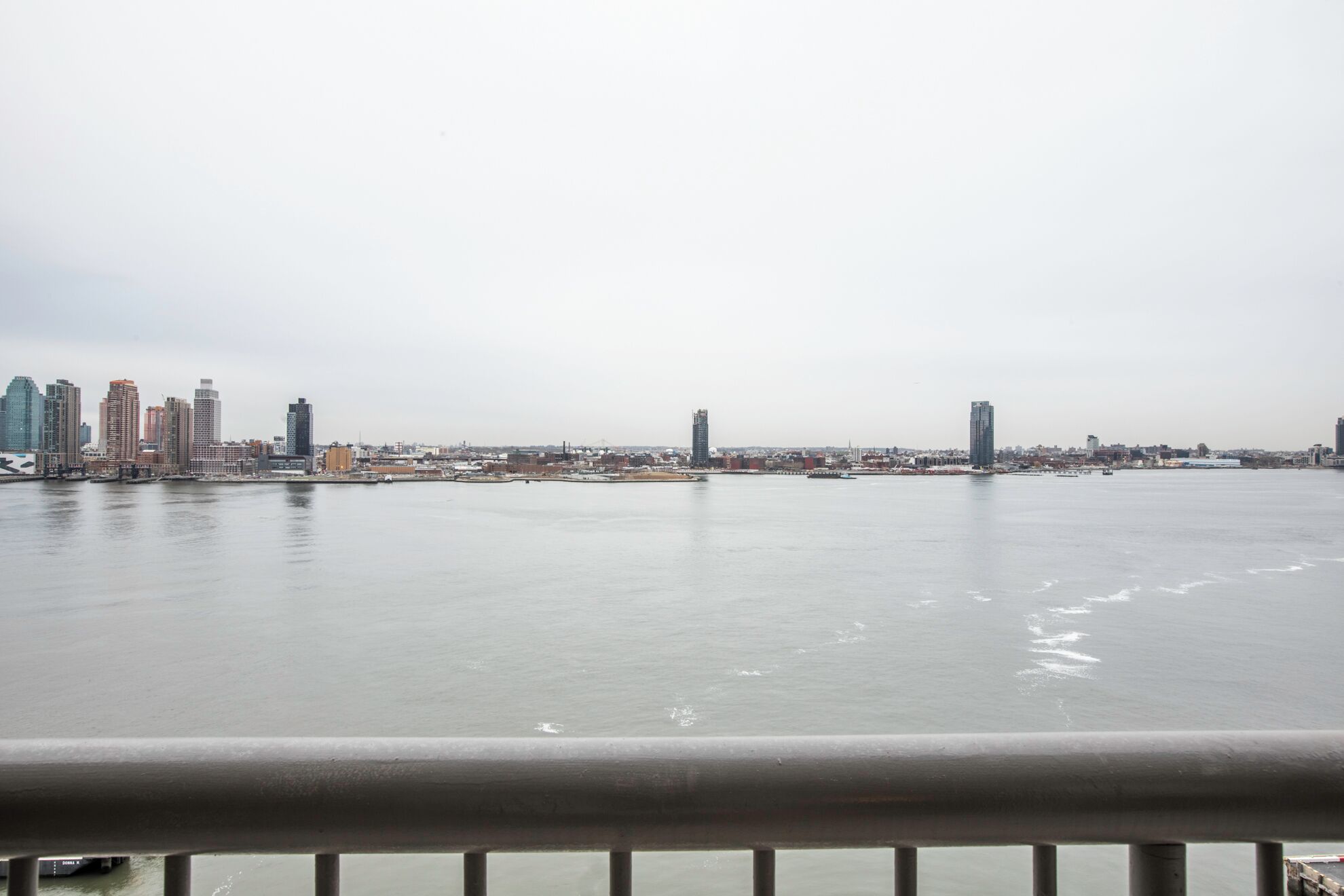 Une vue sur la ville, avec des immeubles de grande hauteur le long du littoral, une rivière paisible et une rambarde métallique au premier plan.