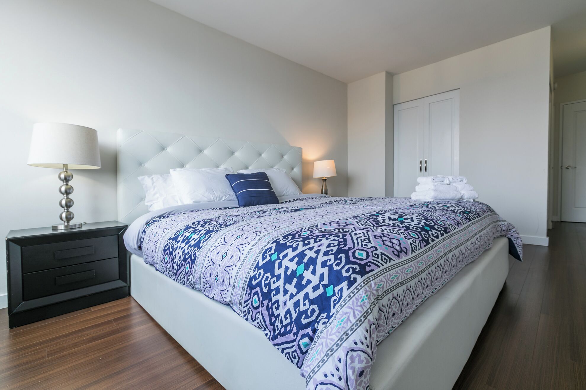 A bedroom with a tufted headboard, a bed with patterned bedding, two bedside tables with lamps, a black nightstand with a drawer, and a white door in the background.