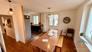 Un grand espace à manger avec une table et des chaises en bois, une cuisine à l’arrière-plan et une horloge au mur.