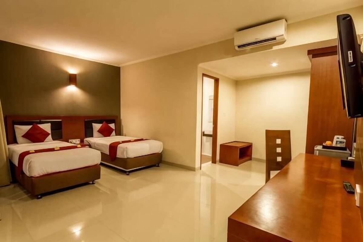 A hotel room with two beds, a wooden desk, a TV, and an air conditioning unit.