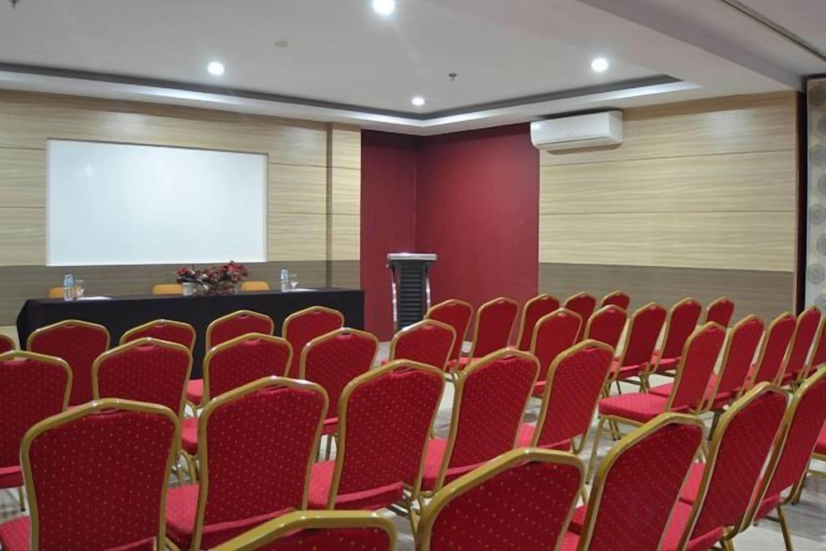 A conference room with rows of red chairs, a podium, and a projector screen.