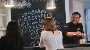 Une personne se tient derrière un comptoir dans un café, et on peut voir, en arrière-plan, un tableau noir sur lequel est écrit : « LE CAFÉ, C’EST UN PALMIER ».