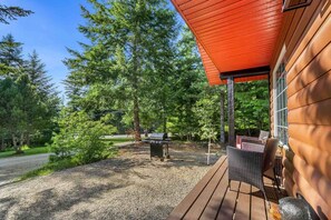 Una terraza de madera con mobiliario exterior, una parrilla y un fondo boscoso.