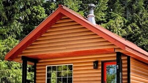 En timmerstuga med ett rött tak, en veranda med två stolar, ett litet bord och en krukväxt.