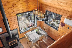 A wooden cabin interior with a dining table, chairs, a black stove, and large windows offering a view of the outdoors.