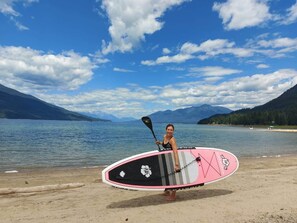 Una persona sostiene una tabla de paddle surf en una playa con montañas al fondo.