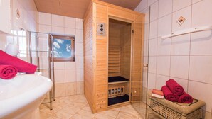 Una stanza sauna con una porta in legno, un box doccia in vetro, un lavandino bianco e un cesto di vimini con asciugamani.