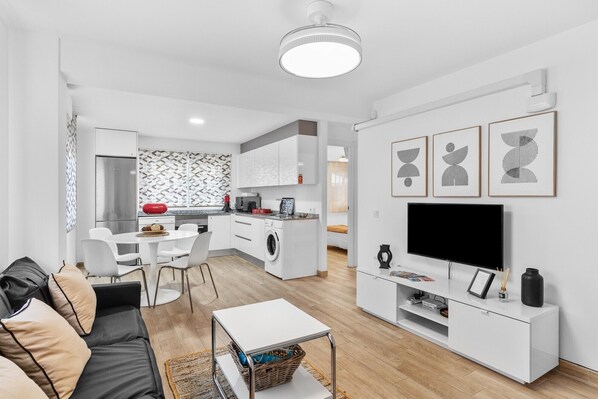 Un salon moderne comprenant un canapé, une table basse, un téléviseur et un coin cuisine avec un lave-linge.