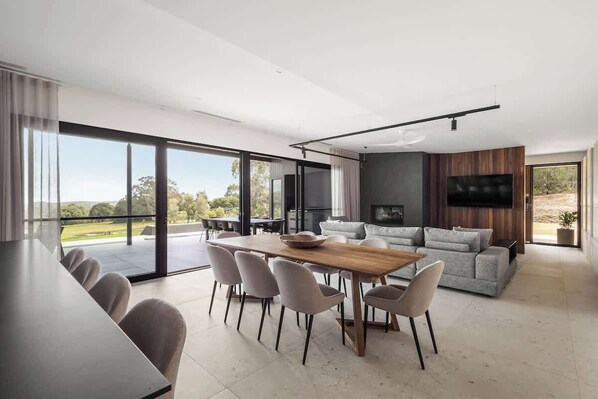 A modern living room with a dining area, large windows, and a flat-screen TV.