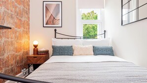 Un dormitorio con una cama, una mesita de noche, una lámpara, un cuadro enmarco y una ventana que ofrece vistas al verdor.