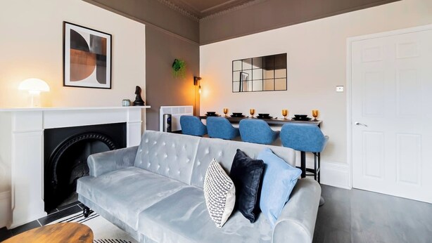 Una sala de estar moderna con un sofá gris, una chimenea, un comedor con sillas azules y una puerta blanca.