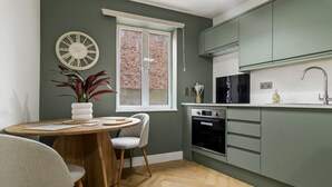A modern kitchen with a dining area, featuring a wooden table, chairs, a potted plant, a clock, and a window with a view of a brick wall. - Pippin - BRAND NEW stylish studio apartment in the hub of Worcester City Centre (Worcestershire)