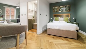 A bedroom with a bed, bedside tables, a nightstand, a window, and a door leading to a bathroom. - Pippin - BRAND NEW stylish studio apartment in the hub of Worcester City Centre (Worcestershire)