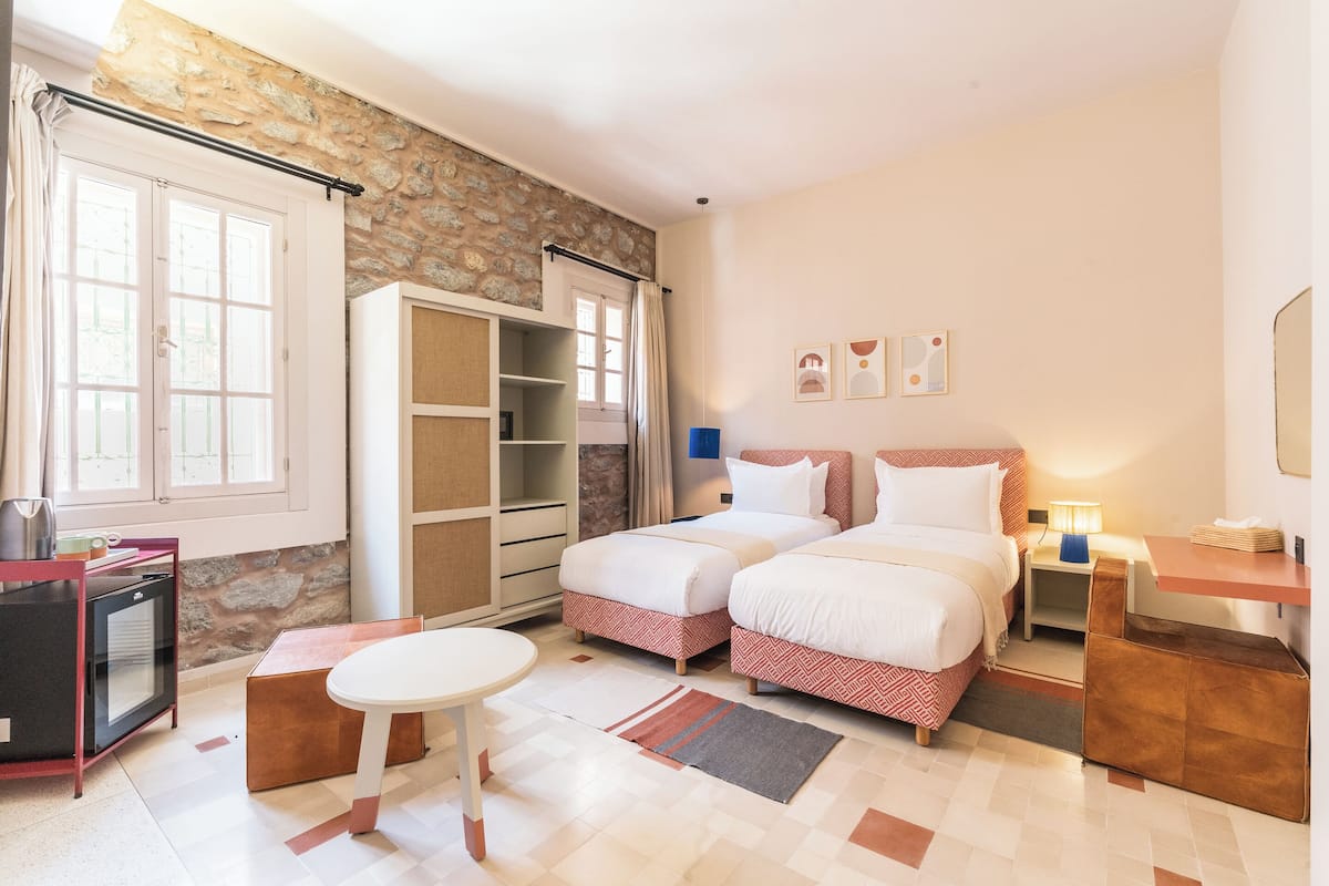 A bedroom with two beds, a small table, a wardrobe, and a kitchenette.