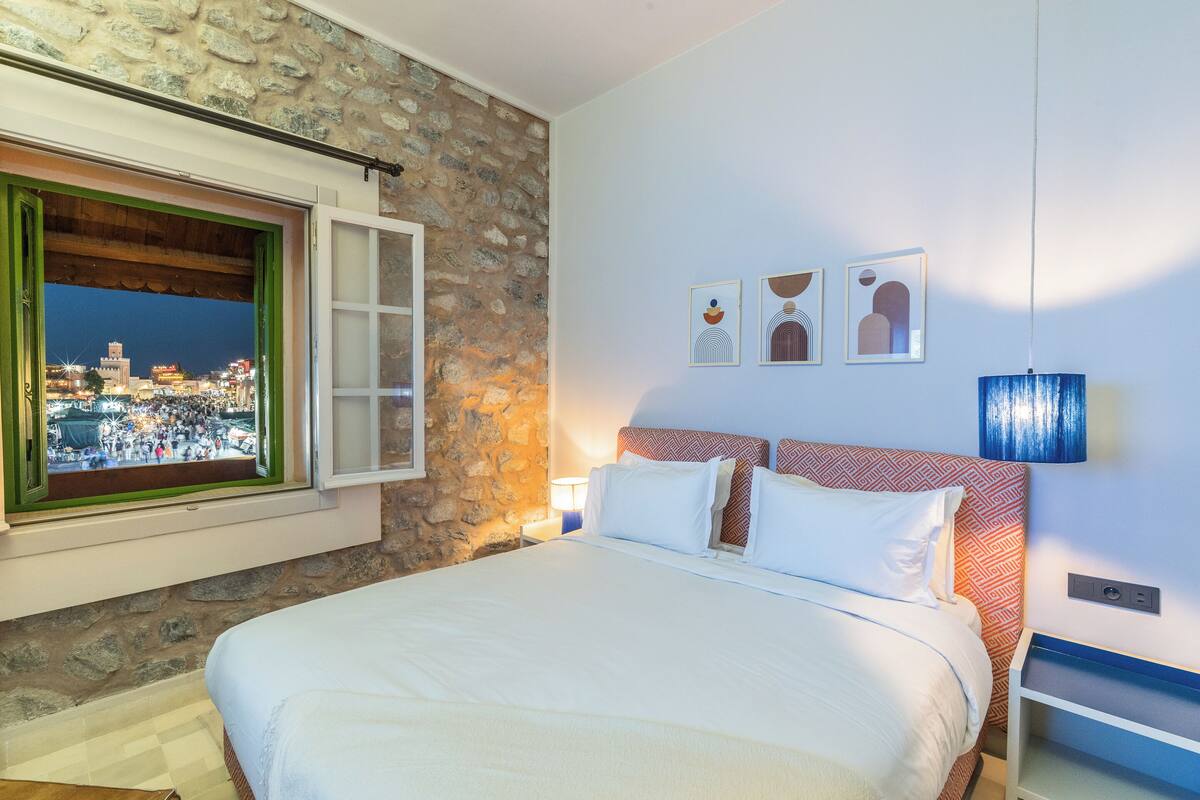 A bedroom with a stone wall, a bed with white linens, a window with a view of a cityscape, and a nightstand with a lamp.