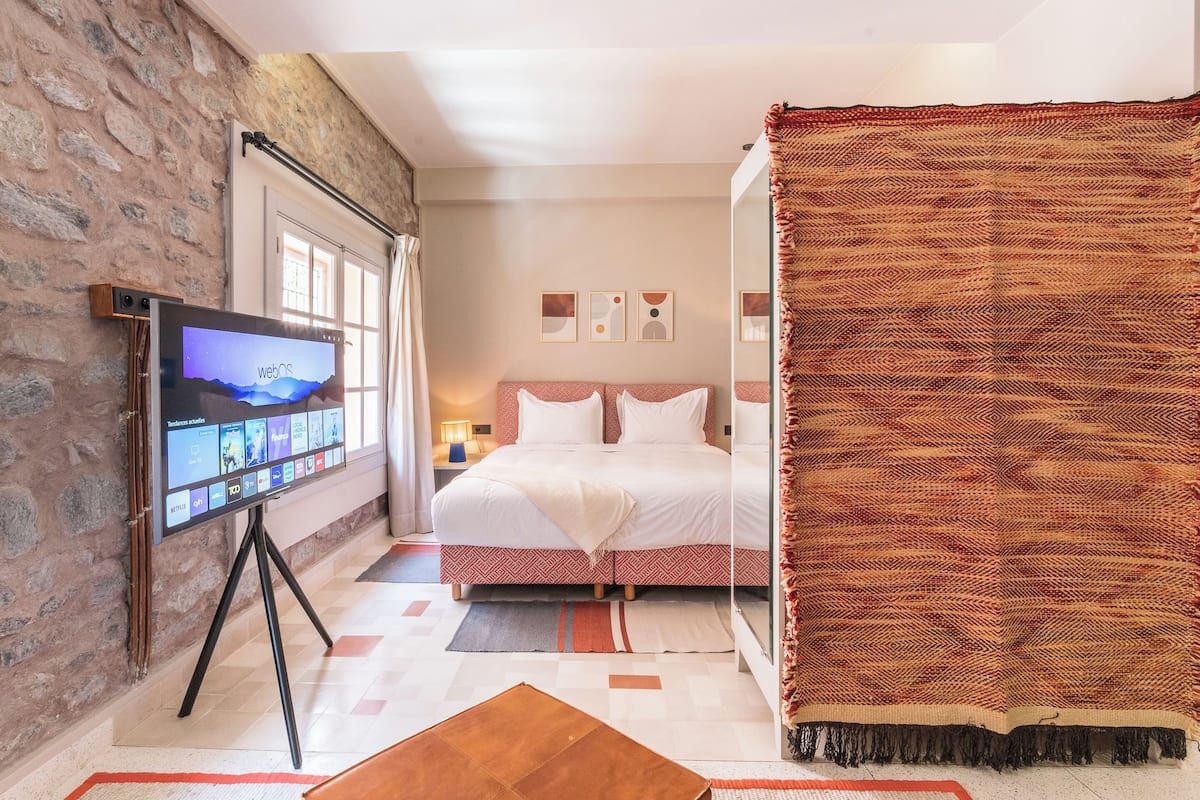A bedroom with a bed, a TV on a stand, a window, and a wall with a stone texture.