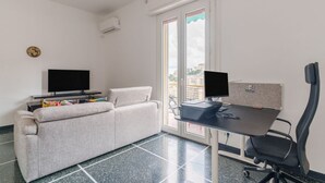 Una habitación con sofá, un escritorio con dos monitores, una silla y una ventana con vista a edificios.