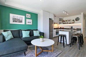 A modern living room with a dark sofa, a round coffee table, and a kitchen area with a bar and stools. - Castle Rock by ITC Hospitality (Cape Town)