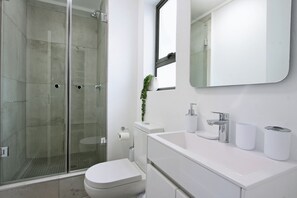 A modern bathroom with a glass shower enclosure, a toilet, a sink with a mirror above, and a small window. - Castle Rock by ITC Hospitality (Cape Town)