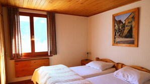 Ein Zimmer mit zwei Betten, Holzboden, einem Fenster mit Vorhängen, einem Nachttisch und einem Bild an der Wand.