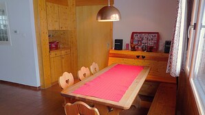 Ein Essbereich mit Holztisch und -stühlen, einer roten Tischdecke, einer hängenden Pendelleuchte und einer Küchenzeile mit Holzschränken und einem Kühlschrank.