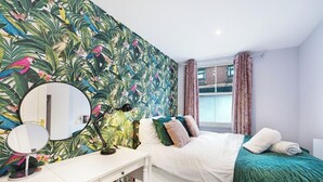 Quarto com cama, mesa de cabeceira com espelho, escrivaninha, janela com cortinas e papel de parede tropical.