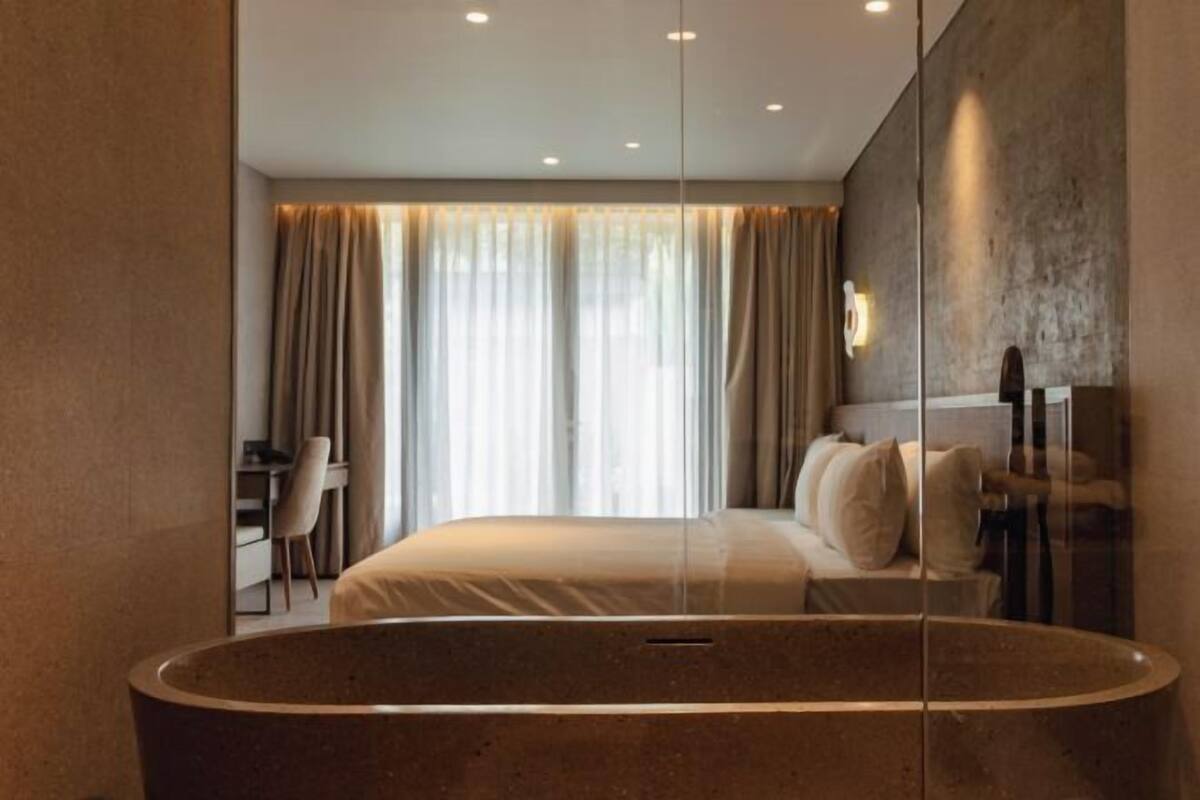 Een moderne hotelkamer met een groot bad, een bed met veel kussens, een bureau en een groot raam met gordijnen.