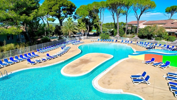 Une piscine bordée de chaises longues bleues et d’une zone de sable, le tout entouré d’arbres et de bâtiments.