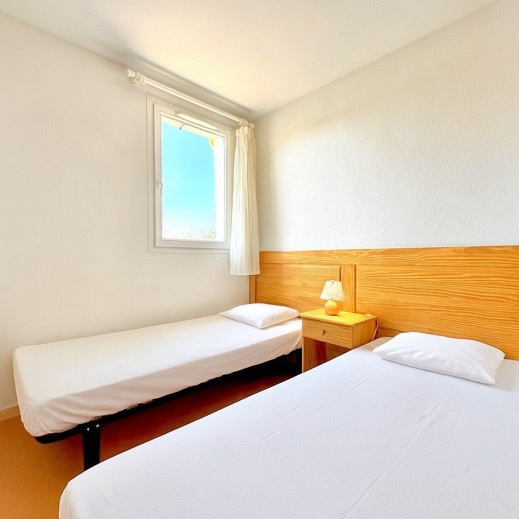 A hotel room with two single beds, a wooden headboard, a bedside table with a lamp, and a window with curtains.