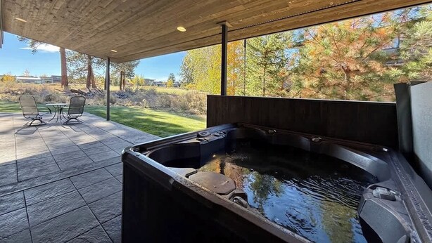 Un espace extérieur aménagé avec un jacuzzi, un plafond en bois, une table avec des chaises et une vue sur les arbres et l’herbe.