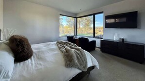 Une chambre moderne avec un grand lit, un téléviseur à écran plat, une console sombre et une vue sur les arbres à travers de grandes fenêtres.