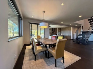 A modern dining area with a wooden table and chairs, a hanging light fixture, and a view of the kitchen and staircase.