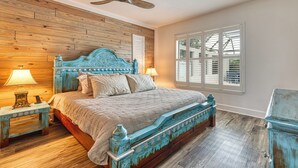 Un dormitorio con cabecera de madera, cama con cubrecama color beige, mesita de noche con lámpara y ventana con persianas.