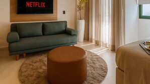 A modern living room with a green sofa, a round ottoman, a flat-screen TV displaying "NETFLIX", a potted plant, and a large window with curtains.