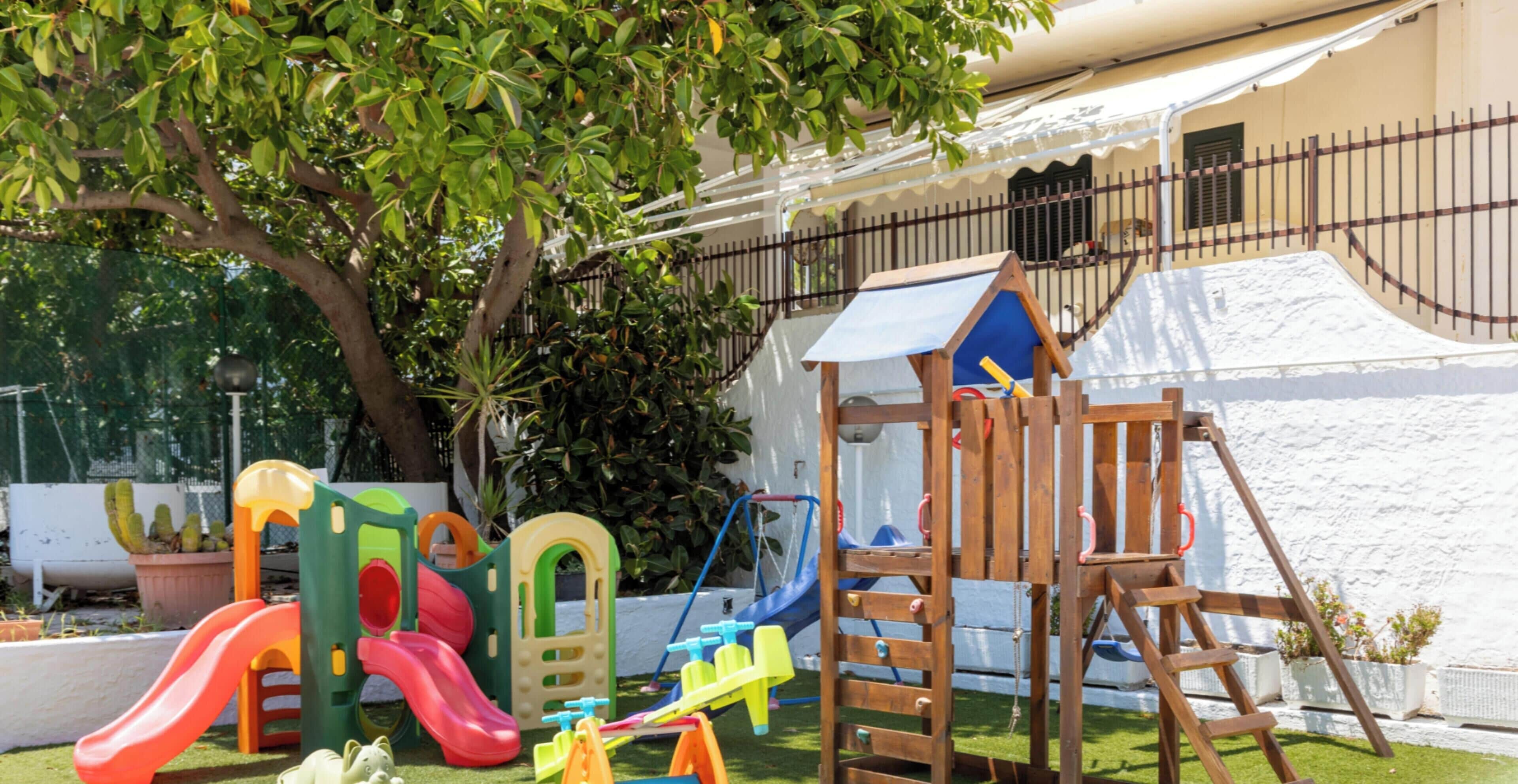 A playground with slides and climbing structures, a tree, and a white wall in the background.