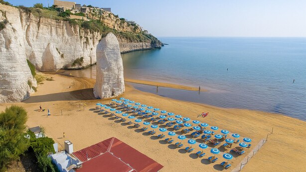 A beach with white cliffs, a row of blue umbrellas, and a red-roofed building. - Hotiday Vieste Centro (Vieste)