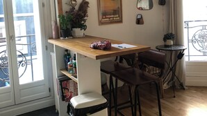 A kitchen with a wooden table, bar stools, a trash bin, a potted plant, and a poster on the wall. - Bright apartment with balcony (Clichy)