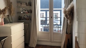 A room with a wooden floor, white furniture, a window with curtains, and a view of buildings outside. - Bright apartment with balcony (Clichy)