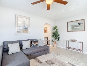 A living room with a grey sectional sofa, patterned cushions, a ceiling fan, a large framed picture, a potted plant, and a console table with a bowl.