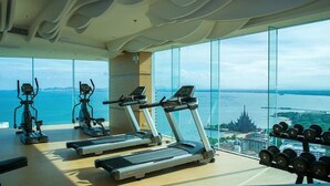 A gym with treadmills, elliptical machines, and dumbbells, overlooking a scenic view of a body of water and a cityscape.
