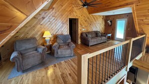 Ein gemütliches Dachzimmer mit Holzwänden, einem Deckenventilator, einem Sofa, einem Sessel und einer Lampe.