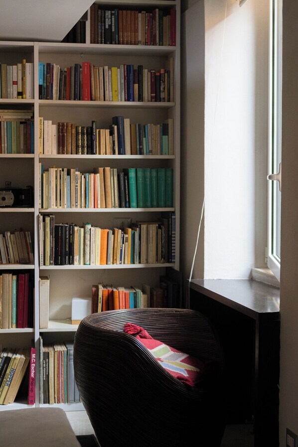 A room with a large bookshelf filled with books, a wicker chair with a blanket, and a window providing natural light.
