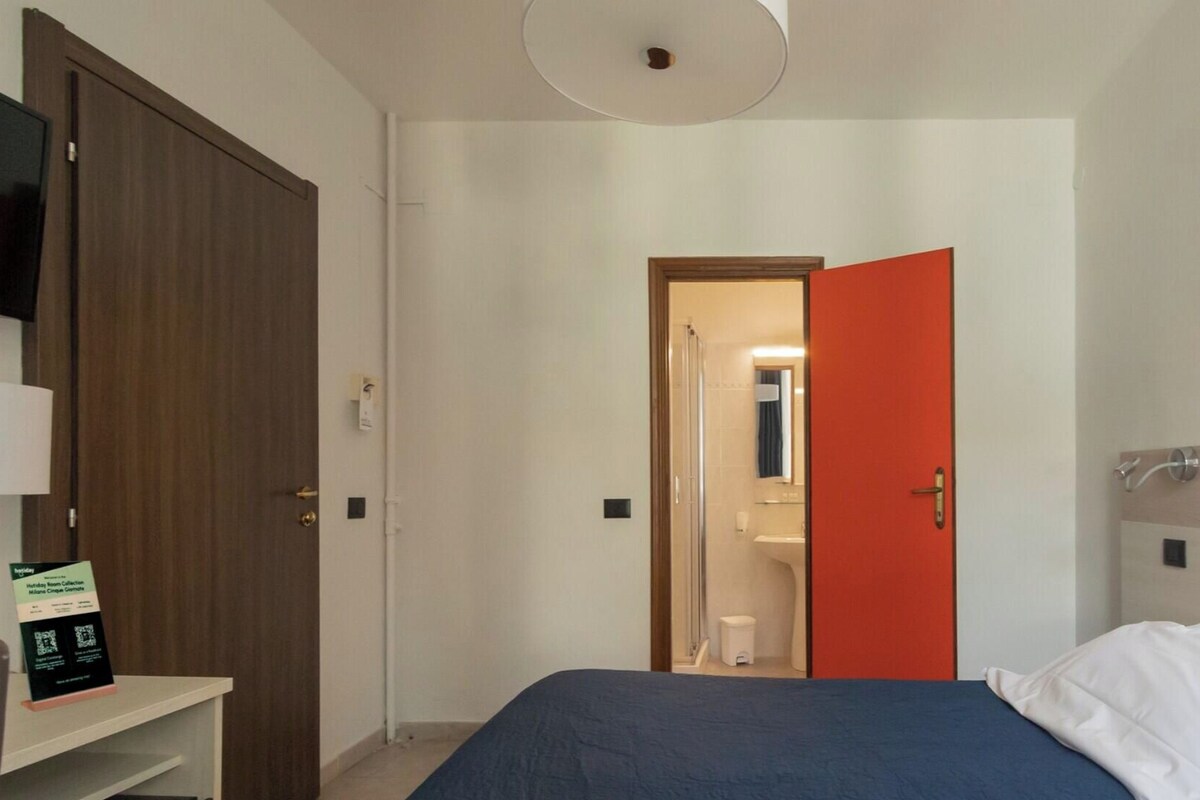 Une chambre d’hôtel comprenant un lit, une télévision, une porte donnant accès à une salle de bain et une œuvre d’art accrochée au mur.