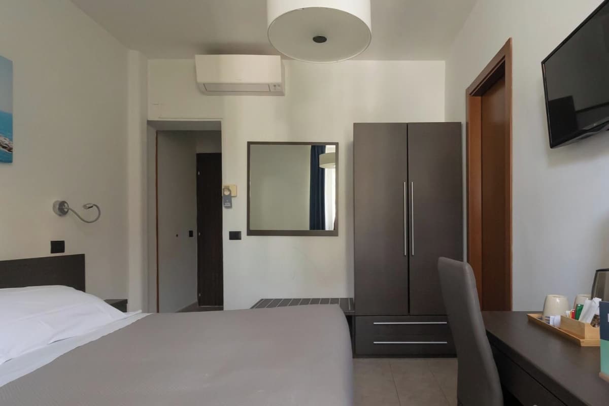 Quarto de hotel com cama, escrivaninha, guarda-roupa, espelho e televisão.