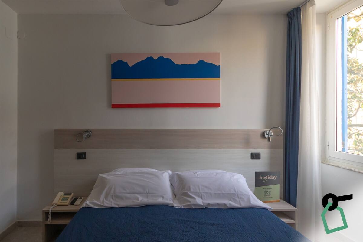 A hotel room with a bed, a bedside table with a phone, a painting on the wall, and a window with curtains.
