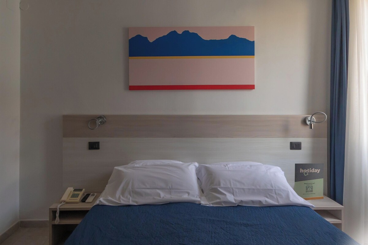 A hotel room with a bed, a bedside table with a phone, a painting on the wall, and a window with curtains.