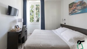 Ein Hotelzimmer mit Bett, Schreibtisch mit Stuhl, Fernseher, Fenster mit Vorhängen und einem Bild an der Wand.
