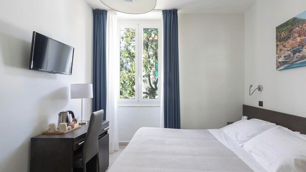 Ein Hotelzimmer mit Bett, Schreibtisch mit Stuhl, Fernseher, Fenster mit Vorhängen und einem Bild an der Wand.